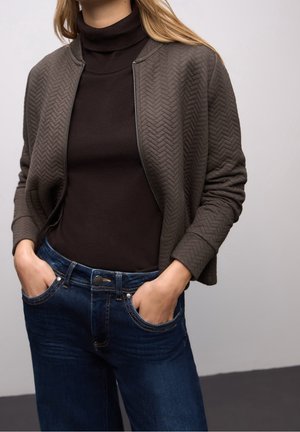 Femme portant une veste matelassée marron foncé sur un pull à col roulé noir et un jean bleu avec les mains dans les poches.