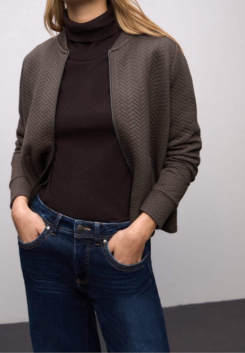 Femme portant une veste matelassée marron foncé sur un pull à col roulé noir et un jean bleu avec les mains dans les poches.