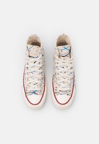 Vysoké tenisky vyrobené z lehkého béžového plátna s tečkovaným vzorem v černé a modré barvě, bílé šňůrky a červené gumové detaily.