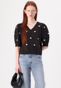 Maglione nero con motivo a cuori bianchi, scollo a V, maniche corte a sbuffo, abbinato a jeans azzurri chiari e una borsa nera annodata.