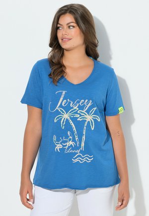 Frau mit braunen Haaren, die ein blaues "Jersey Island" T-Shirt mit Palmen, Krabbe und Wellen trägt, kombiniert mit weißen Hosen, schaut nach rechts.