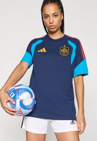 Γυναίκα κρατώντας μια μπλε και λευκή ποδοσφαιρική μπάλα, φορώντας μια ναυτική μπλούζα ποδοσφαίρου της Ισπανίας με κόκκινες και κίτρινες ρίγες και λευκές βερμούδες Adidas.