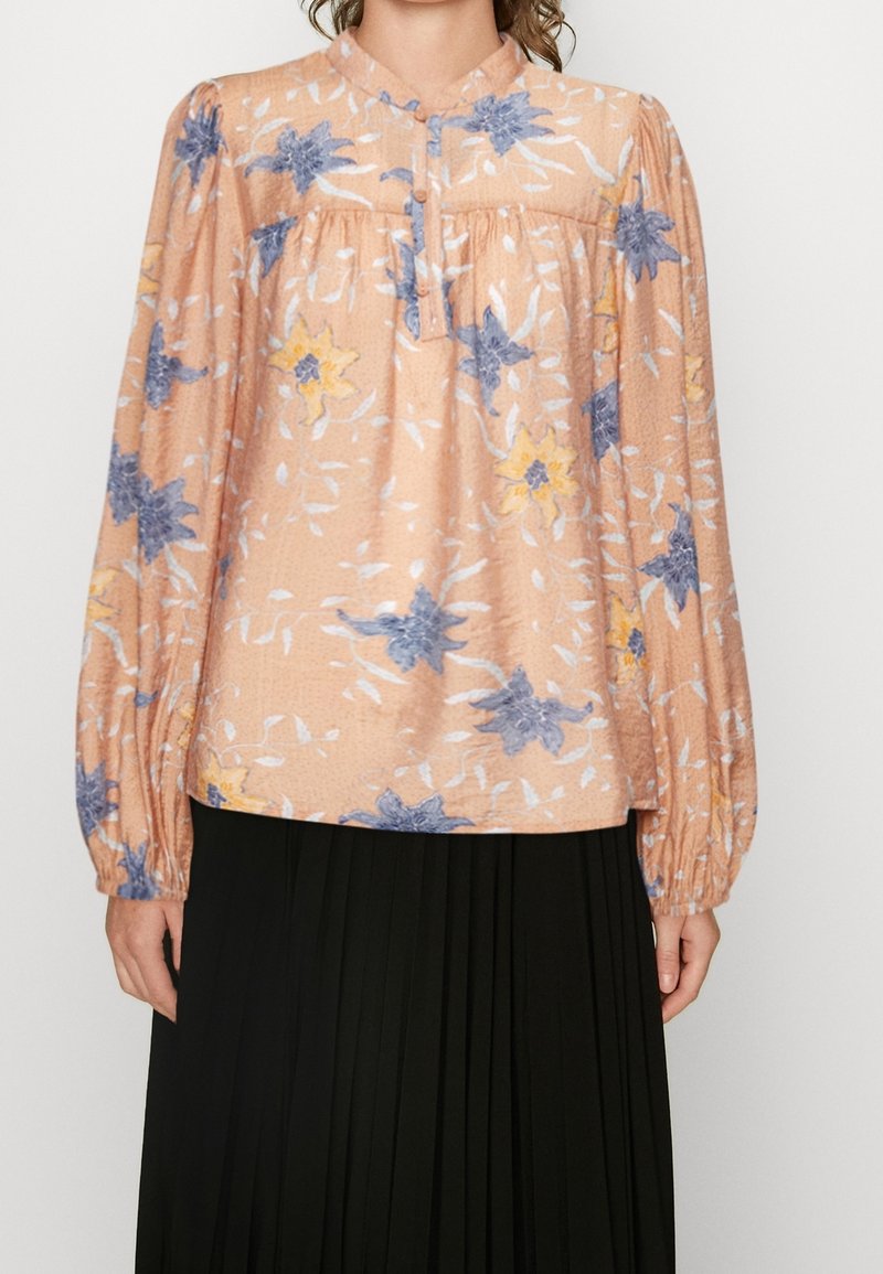 Femme portant un chemisier floral pêche avec des fleurs bleues et jaunes et des manches longues bouffantes, associé à une jupe plissée noire.