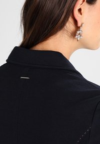 Woman’s side profile showing long brown hair, crystal and gold drop earring, and black blazer collar with small metal tag.