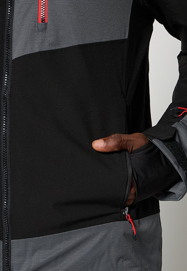 Mano con chaqueta negra y gris con tiradores de cremallera rojos, pulgar insertado en el bolsillo lateral, primer plano del torso y el brazo.