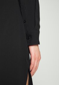 Gros plan d'une main à côté d'un vêtement noir à manches longues avec poignets boutonnés et détails avec fente latérale.