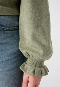 Trui in olijfgroen met pofmouwen, een gestructureerde afwerking en gerimpelde ruches bij de manchetten. Gecombineerd met denim, wat een casual stijl laat zien.