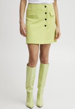 Limegrön minikjol med framfickor, dekorerad med fem svarta knappar och rå nederkant, kombinerad med knähöga, texturerade matchande gröna stövlar.