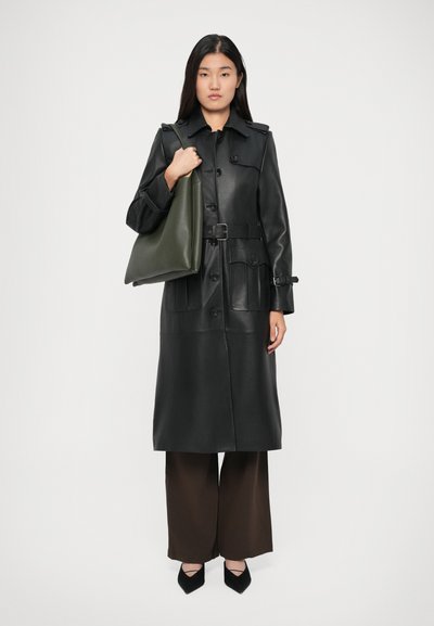 Manteau trench en cuir noir avec des boutons sur le devant, deux poches poitrine et une ceinture à la taille. Le modèle tient un sac fourre-tout en cuir vert, associé à un pantalon large marron.