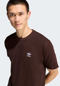 Hnedé bavlnené tričko s okrúhlym výstrihom a krátkymi rukávmi, s bielym logom Adidas na ľavej strane hrudníka. Hladká textúra.