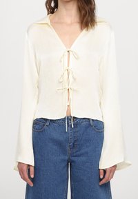 Lys cremefarvet bluse med lange ærmer, bindesløjfe foran og krave; blødt, tekstureret stof; kombineret med løse blå denimjeans.