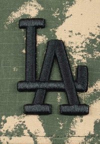 Black embroidered "LA" logo on a green and beige camouflage fabric, featuring textured stitching and defined letter shapes.
