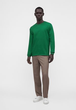 Homme debout portant une chemise verte à manches longues, un pantalon beige et des baskets blanches sur un fond gris uni.