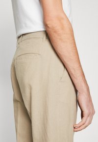Pantalons en lin beige au coupe décontractée, avec une poche arrière et un tissu texturé. La taille est plate avec une finition subtile.