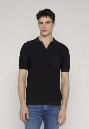 Junger Mann mit kurzen lockigen Haaren, der ein schwarzes kurzärmeliges Hemd mit Kragen und blaue Jeans vor einem schlichten hellen Hintergrund trägt.