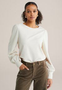 Mlada ženska s kodrastimi lasmi nosi belo bluzo z dolgimi rokavi in napihnjenimi rokavi ter rjave kordurove hlače, samozavestno pozira.