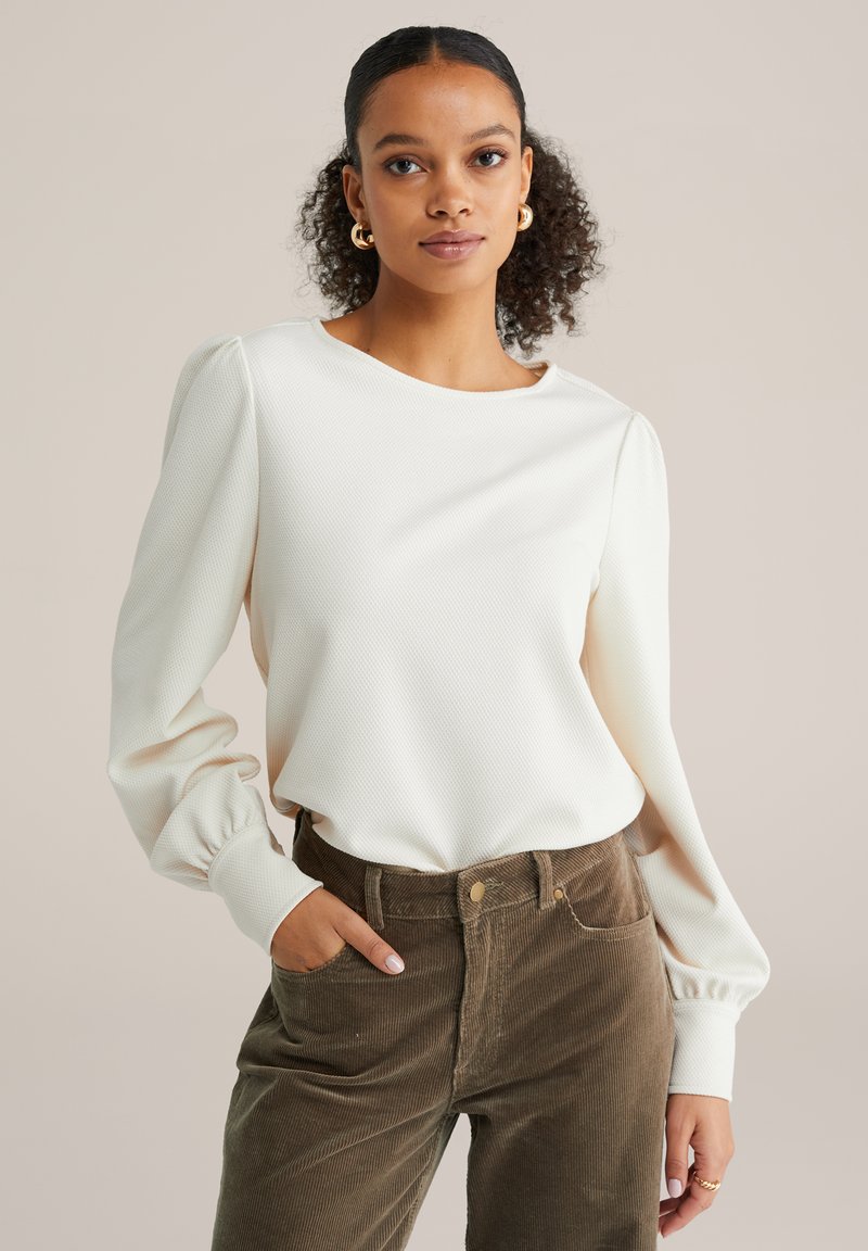 Mlada ženska s kodrastimi lasmi nosi belo bluzo z dolgimi rokavi in napihnjenimi rokavi ter rjave kordurove hlače, samozavestno pozira.