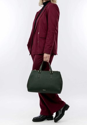 Persona con traje burdeos y botas negras sosteniendo un gran bolso de cuero negro sobre un fondo blanco liso.