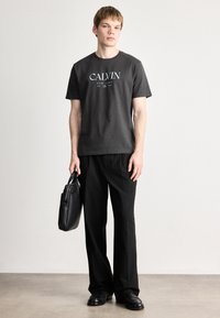 Must musta T-särk valge "CALVIN" trükiga, kombineeritud mustade laia jala pükste ja musta nahkkotiga, neutraalse taustaga.