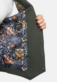 Olivgrüne Jacke mit einem gemusterten, marineblauen Innenfutter, das Blumen- und Paisleymuster zeigt. Das Etikett trägt die Aufschrift "JEFF URBAN TAILORING 52."