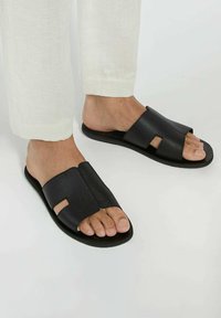Black leather sandals with a textured finish, featuring two wide straps and an open toe design. The sole is flat and made of soft material.