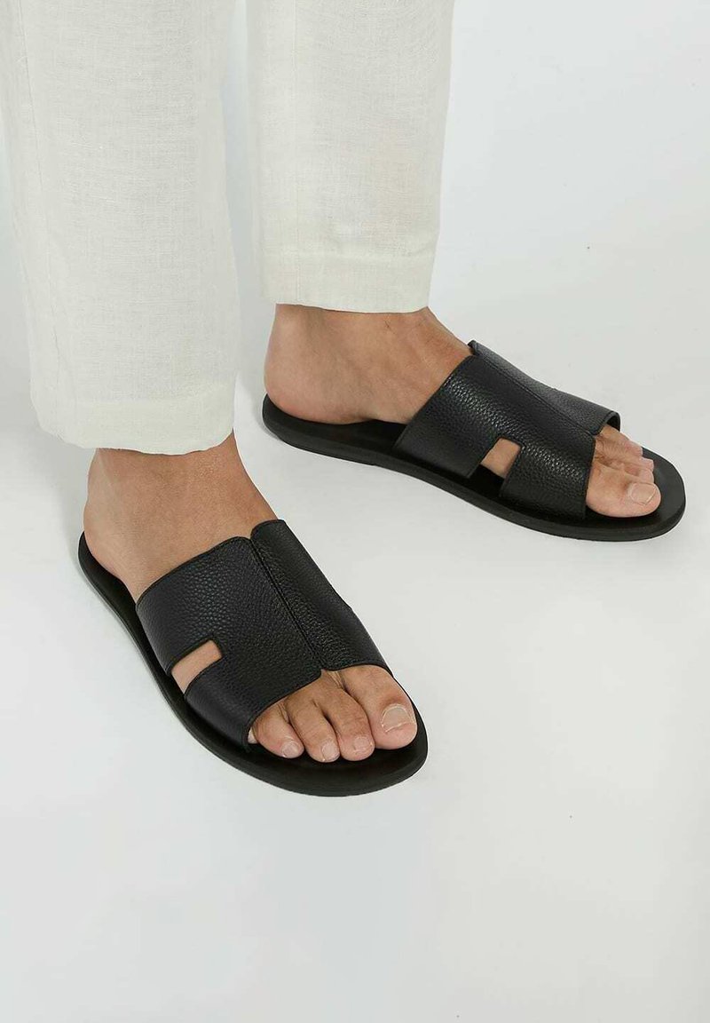 Black leather sandals with a textured finish, featuring two wide straps and an open toe design. The sole is flat and made of soft material.