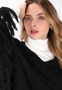 Schwarzer, übergroßer Pullover mit strukturierten Fransenärmel, über einem weißen Rollkragenpullover getragen. Glatte Haut und dezentes Make-up betonen die Gesichtszüge.