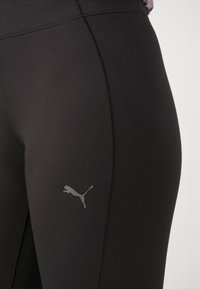 Czarne legginsy sportowe o gładkiej teksturze, z przeszytymi szwami i szarym logo Pumy na lewym udzie.