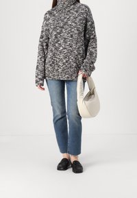 Gestrickter schwarz-weiß gesprenkelter Pullover mit hohem Kragen, kombiniert mit blauen Jeans mit geradem Bein und schwarzen Loafers. Eine weiße Handtasche wird gehalten.
