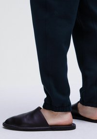 Zapatos negros de cuero tipo slip-on con punta redondeada y suela plana, combinados con pantalones oscuros de corte ajustado con puños elásticos. Textura suave.