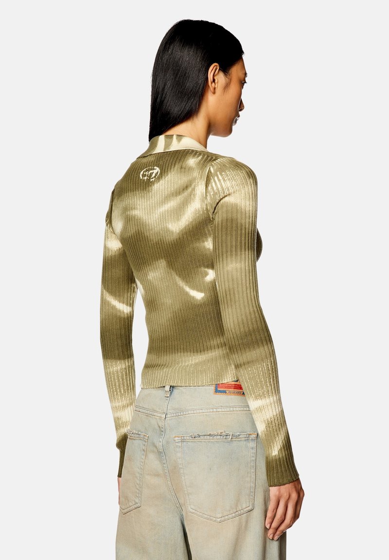 Pull en maille côtelée vert olive avec une coupe ajustée, texture brillante et manches longues, présentant un détail de logo dans le dos. Associé à un jean clair.