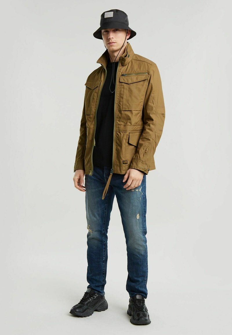 Chaqueta verde oliva con múltiples bolsillos, suéter oscuro debajo, jeans azules desgastados y zapatillas negras. Combinado con un sombrero tipo cubo negro.