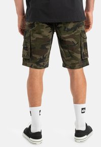 Pantalones cortos cargo con patrón de camuflaje, fabricados en algodón. Cuentan con dos bolsillos laterales, cinturilla negra y un corte holgado. Llevan calcetines blancos y zapatillas negras.