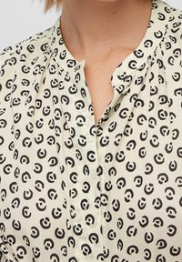Bluzka w kolorze kremowym z czarnym abstrakcyjnym wzorem, z zapięciem na guziki i plisowanymi detalami wzdłuż przodu.