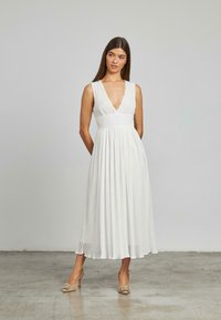 Abito bianco plissè con scollatura a V profonda, vita aderente e gonna fluida. Indossato con sandali con tacco beige caratterizzati da dettagli decorativi.