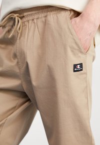 Ljust beige, byxorna med elastisk midja och dragsko. Har sidofickor och en liten Champion-logotyppatch på vänstra låret. Slät textur.