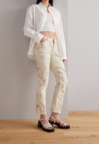 Värgglada jeans med abstrakta mönster i pastellfärger, kombinerade med en vit knäppskjorta och sandaler med blockklack och remmar.