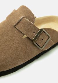 Gros plan d'une chaussure marron en daim à enfiler, avec une bride réglable à boucle métallique et une semelle noire.