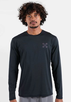 Homme aux cheveux bouclés portant un t-shirt noir à manches longues avec un petit logo géométrique violet sur la poitrine gauche.