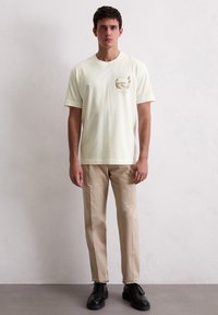 Bărbat îmbrăcat cu un tricou grafic de culoare albă murdară, pantaloni bej și pantofi negri cu șireturi, stând în fața unui zid simplu de culoare gri deschis.