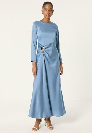Femme portant une robe bleue en satin à manches longues et longueur au sol, avec une boucle décorative dorée à la taille, associée à des sandales beiges à talons hauts.