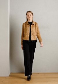 Tan suede jacket with a collared neckline, front snap closures, and two chest pockets, worn over a black top and black trousers.