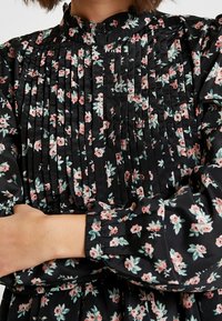 Abito nero floreale con piccoli fiori rosa e verdi, design plissettato, colletto alto e maniche lunghe con polsini arricciati.