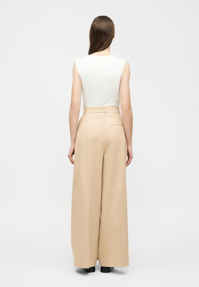 Woman with long brown hair wearing a white sleeveless top and high-waisted wide-leg beige trousers, standing facing away on white background.