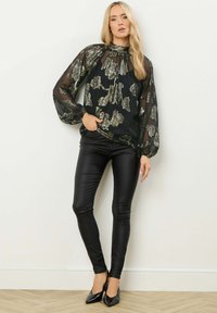 Blusa nera trasparente con motivo floreale metallico, scollatura a sbuffo e maniche lunghe a palloncino, abbinata a pantaloni in pelle nera aderenti e scarpe con tacco a punta.