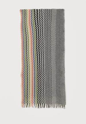 Prostokątny kawałek tkaniny z zygzakowatymi paskami w kolorach czarnym, białym, czerwonym, niebieskim, zielonym i pomarańczowym, z frędzlami na jednym krótkim brzegu.