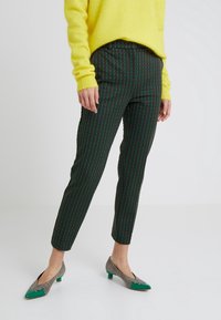 Kvinna iklädd rutiga slim-fit byxor, gröna och bruna mönstrade lågklackade skor, samt en ljusgul tröja, stående på ett vitt golv.