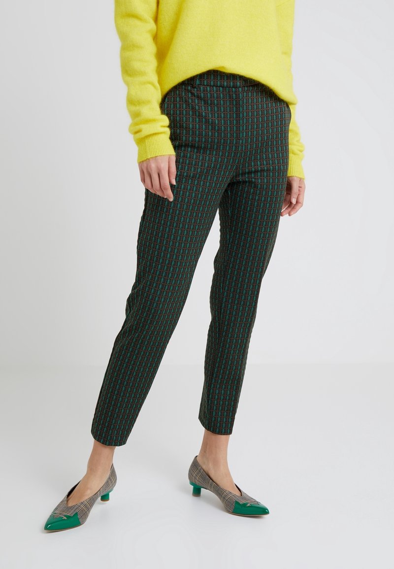 Kvinna iklädd rutiga slim-fit byxor, gröna och bruna mönstrade lågklackade skor, samt en ljusgul tröja, stående på ett vitt golv.