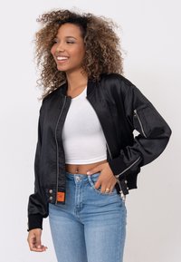 Zwarte satin bomberjack met ritssluitingen en geribbelde manchetten, gedragen over een wit fitted top en gecombineerd met hoge, lichtblauwe jeans.