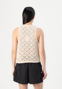 Top senza maniche lavorato a maglia beige con un motivo a rombi, scollatura a coste e trama aperta. Indossato sopra dei pantaloni corti neri.